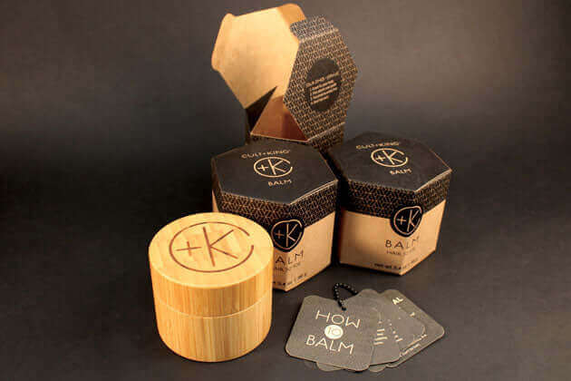 Three hexagonal Cult and King BALM | Hair to Toe containers with black and brown designs are shown next to a wooden jar and black product cards. These moisturizing balms from Cult and King feature organic botanical ingredients on a dark background.