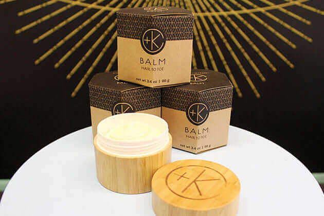 A small open jar of Cult and King BALM | Hair to Toe with a wooden lid is displayed before three matching boxes on a white surface, set against a black and gold background. Perfect for nourishing skin and hair.