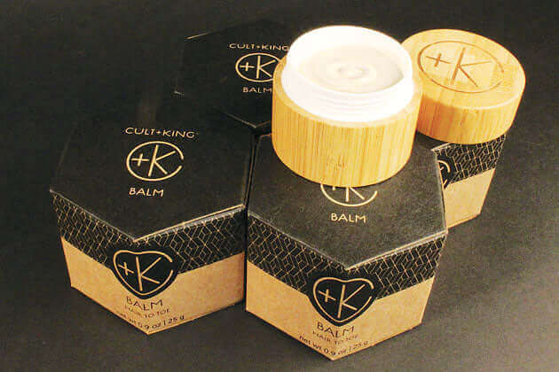 Three hexagonal black and brown containers of Cult and King BALM | Hair to Toe by Cult and King are displayed, two closed and one open showing white balm. Wood lids add a natural look, highlighting its blend of organic botanicals for skin and hair care.