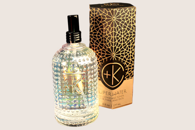 A clear, textured glass bottle labeled "+K" holds Cult and King SUPER WATER Antioxidant Toner for Hair and Skin, shown beside a matching black and gold box with a geometric pattern, highlighting its vegan hydration benefits.