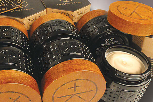Several black jars with textured sides and wooden lids labeled “Cult and King VEGAN BALM | Manifest Moisture for Hair Styling and Face” are stacked, one open to reveal creamy balm inside. Brown Cult and King boxes appear in the background.