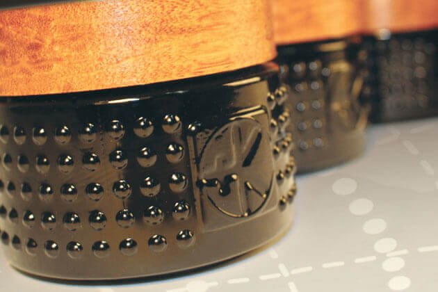 Close-up of Cult and King VEGAN BALM, a cylindrical hair styling tool with a textured black base featuring raised dots and an embossed Cult and King logo, plus a smooth wood-toned top. It rests on a faintly grid-patterned surface.