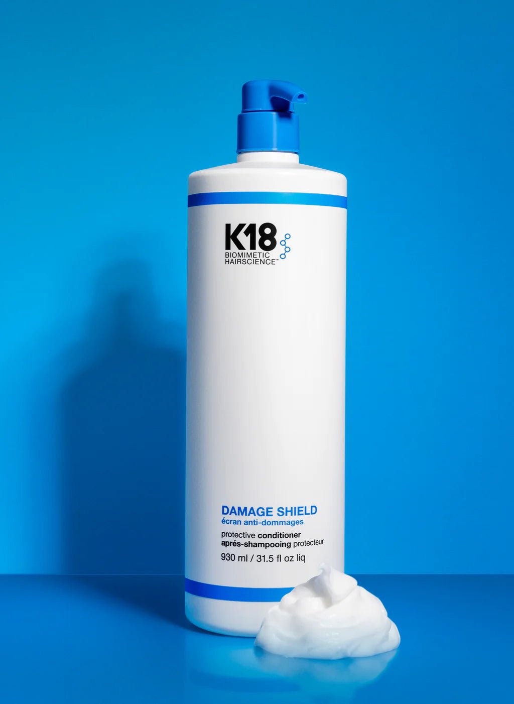 A large white bottle of K18 Hair Repair K18 Damage Shield pH Protective Conditioner with a blue pump on a blue background, next to a dollop of conditioner, emphasizing hair health and damage protection.