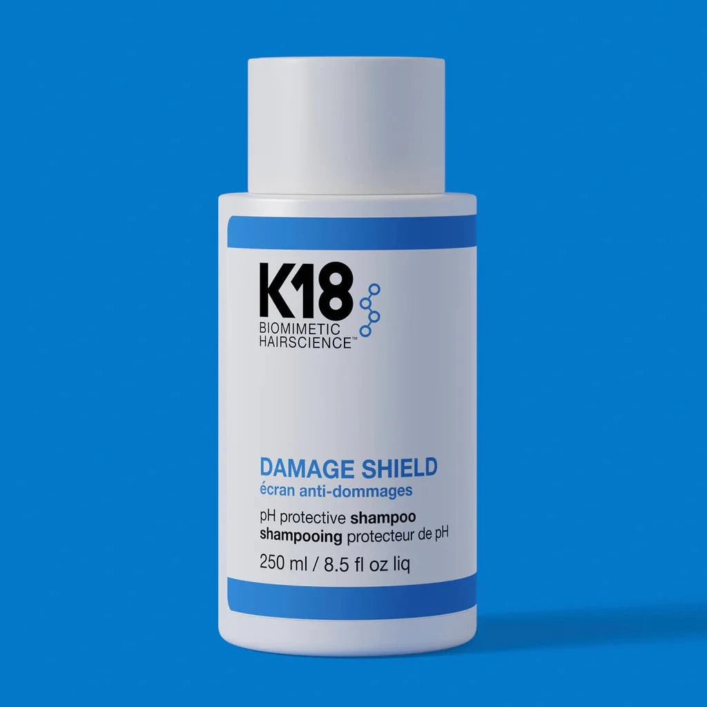 A bottle of K18 Hair Repair DAMAGE SHIELD pH Protective Shampoo (multiple sizes) with K18PEPTIDE™ is displayed against a blue background, featuring a circular label that reads "new name! same formula.