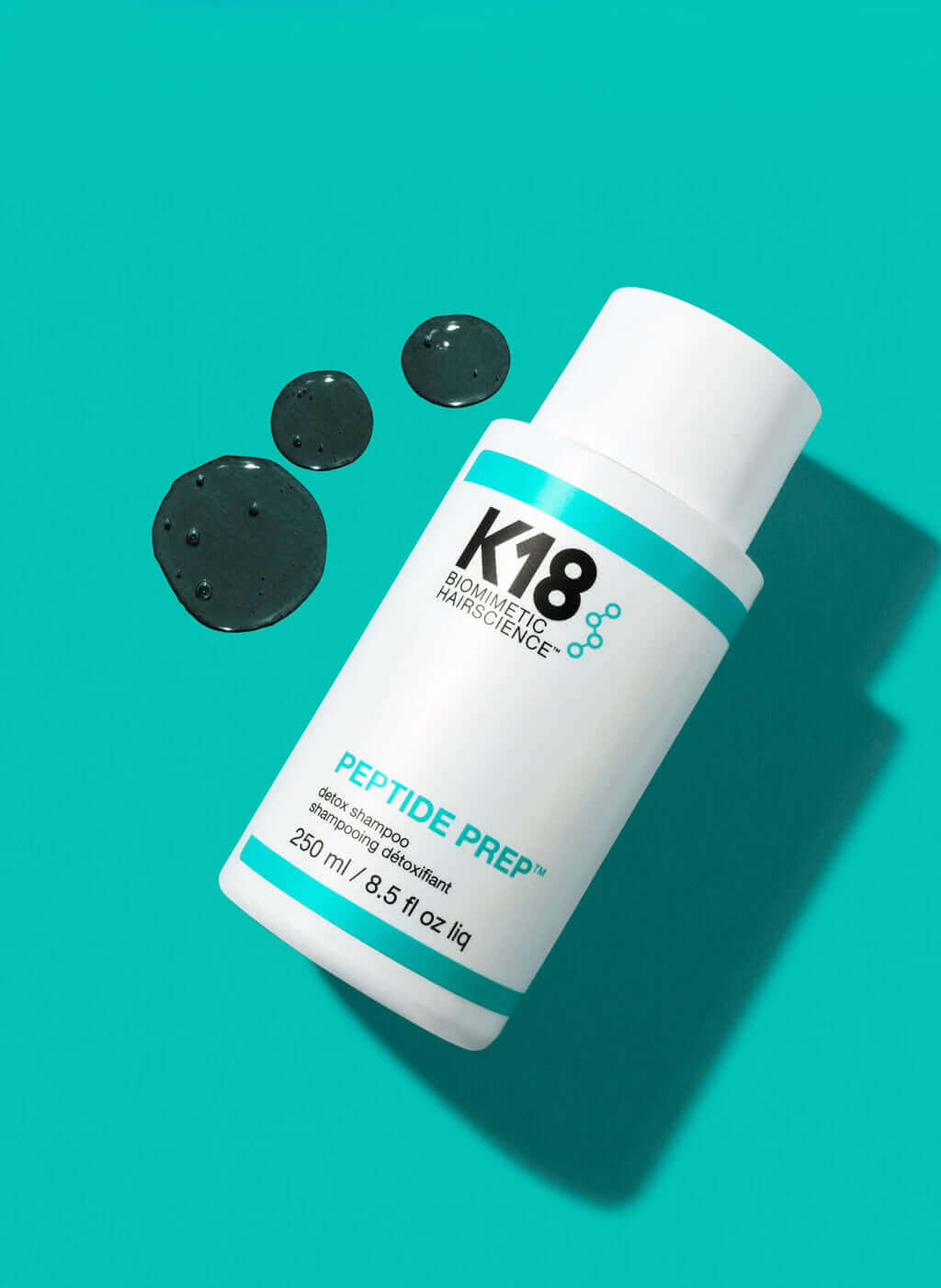A white K18 Hair Repair PEPTIDE PREP Detox Shampoo bottle (250ml/8.5 fl oz) rests on a teal surface beside three drops of clear liquid, highlighting its deep-cleansing formula for hair follicles.
