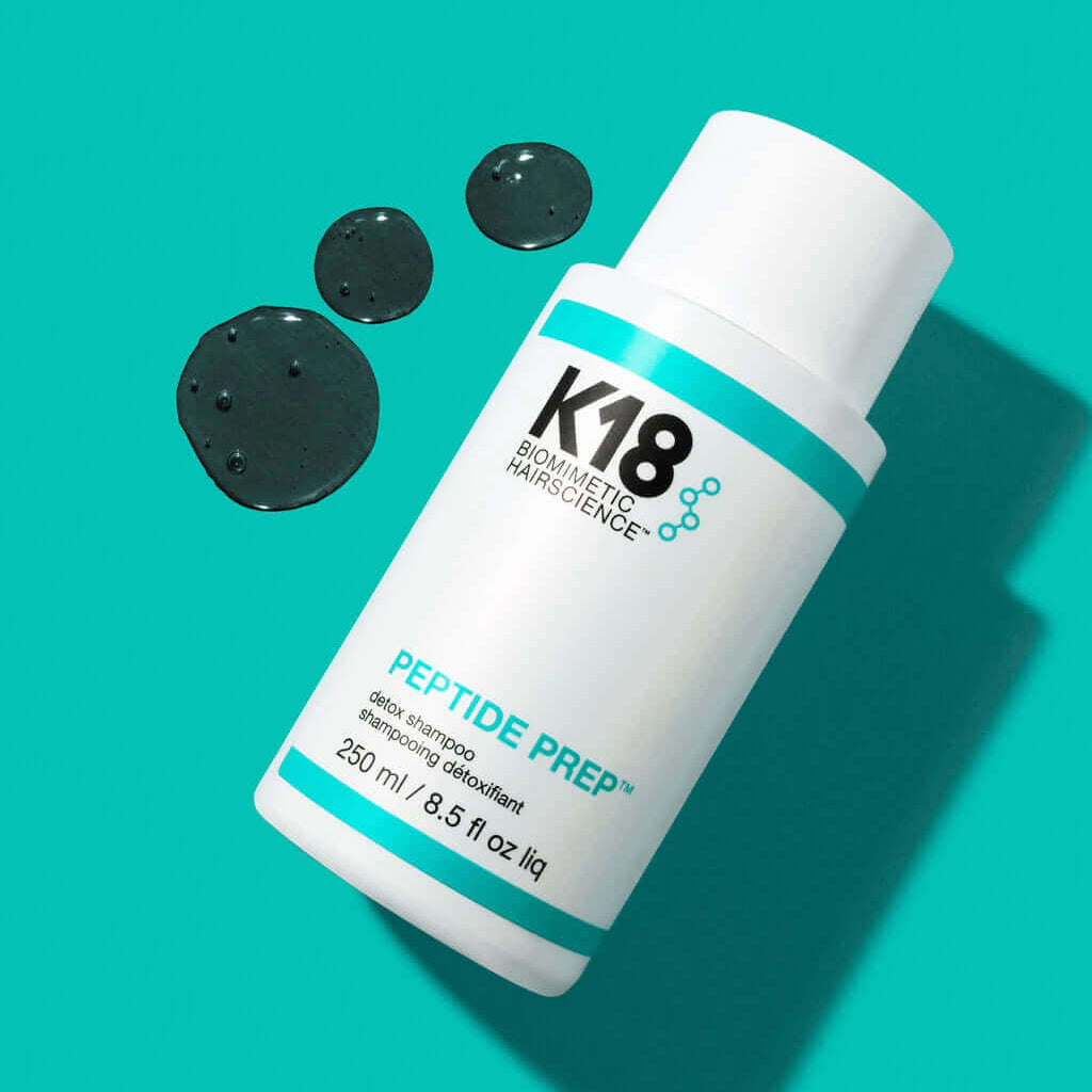 A white K18 Hair Repair PEPTIDE PREP Detox Shampoo bottle (250ml/8.5 fl oz) rests on a teal surface beside three drops of clear liquid, highlighting its deep-cleansing formula for hair follicles.