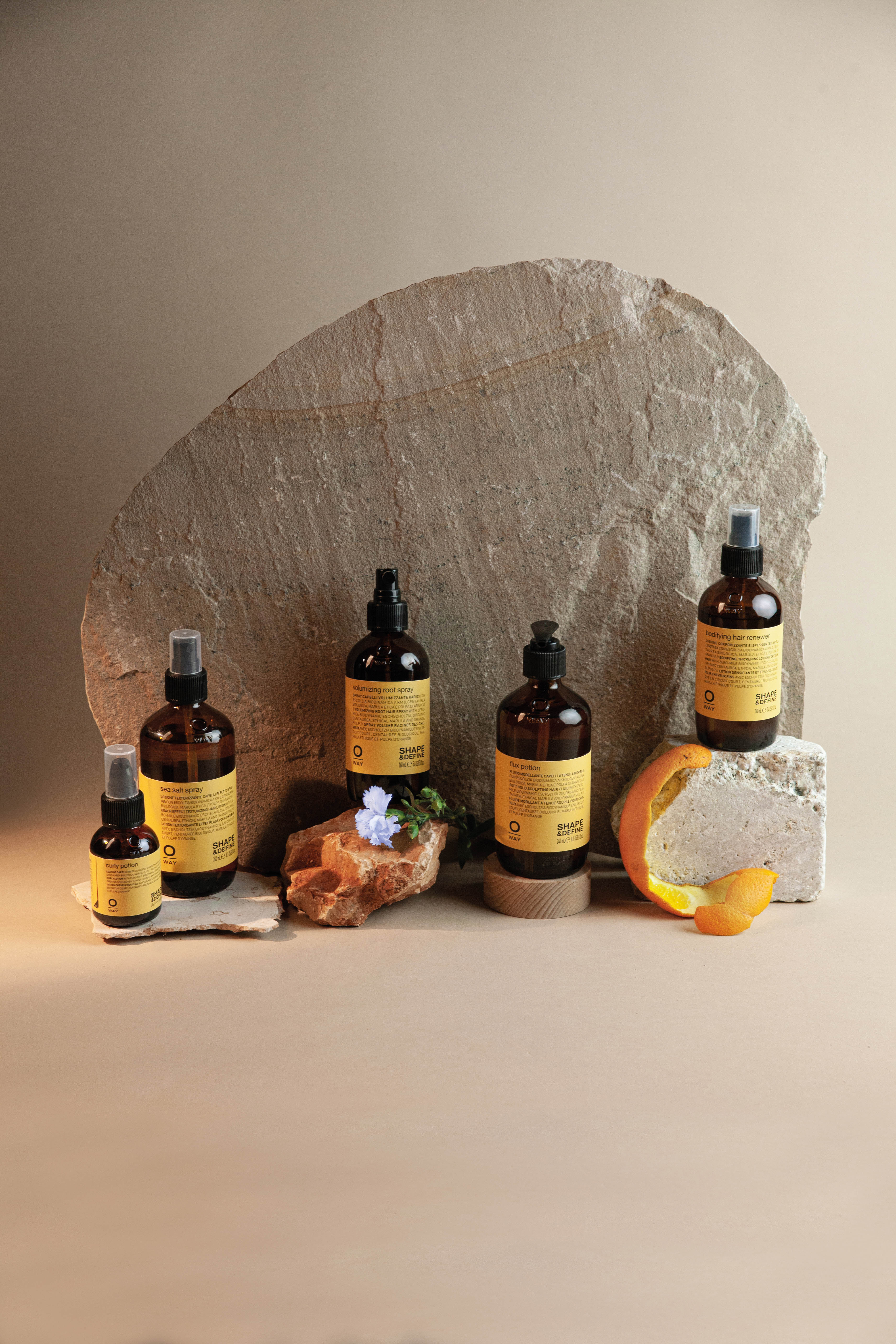 Five amber-colored bottles with yellow labels are arranged on stones, alongside a slice of orange peel and a small blue flower, set against a neutral background.