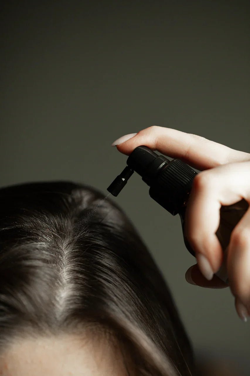 A close up of a hair part and someone spraying product onto the scalp with a grey background.