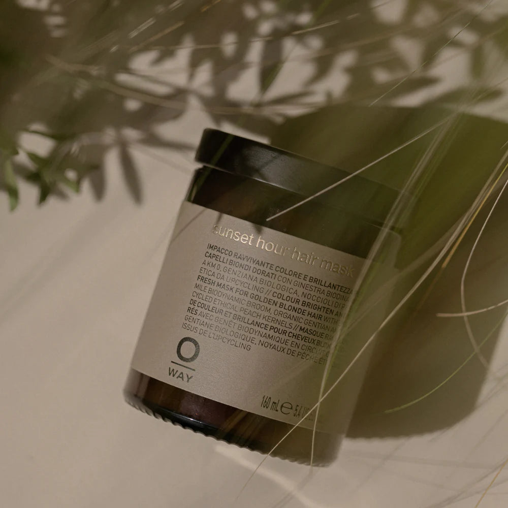 A jar of Oway Sunset Hour Hair Mask | Radiance-Boosting Mask for Golden Blonde Hair sits on a neutral surface, partially shaded by foliage.