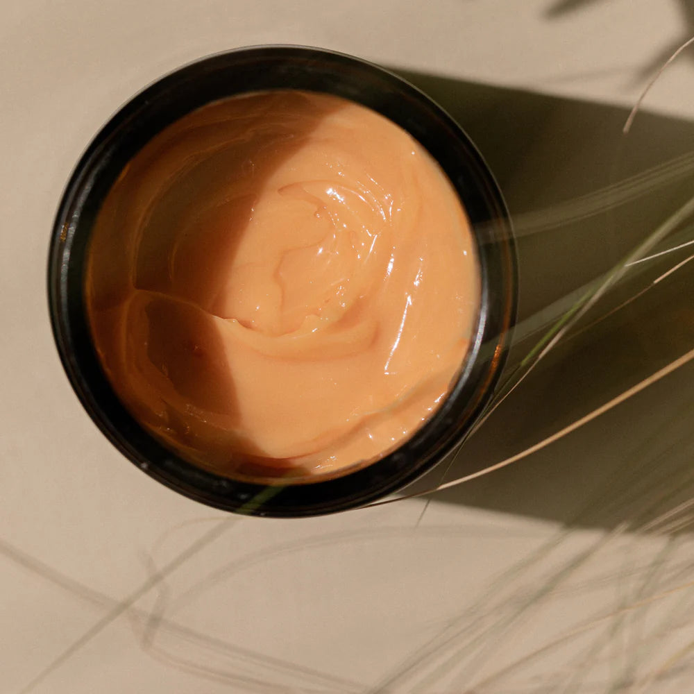 An open black container of Oway Sunset Hour Hair Mask | Radiance-Boosting Mask for Golden Blonde Hair sits on a light surface, peach-colored formula visible inside, with plant shadows partially cast over it.