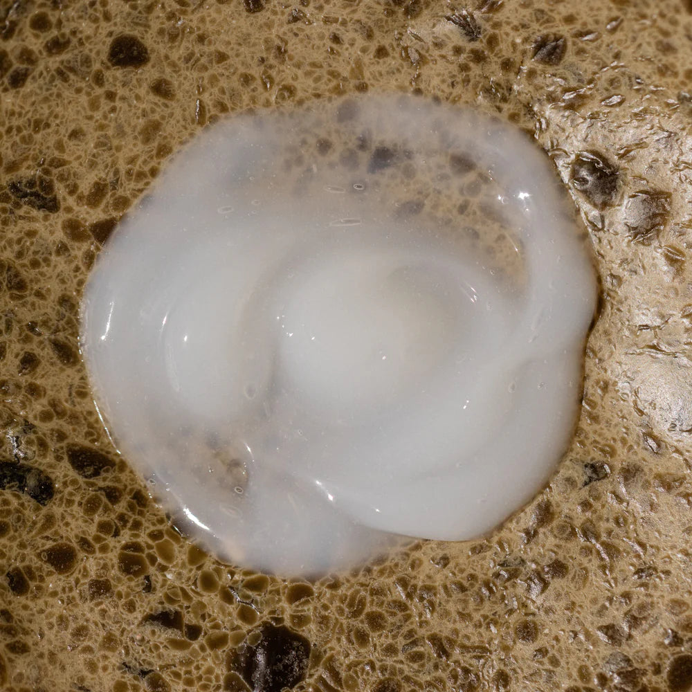 A small puddle of Oway Enriching Hair Conditioner’s creamy, plant-based formula sits on a brown, speckled textured surface.