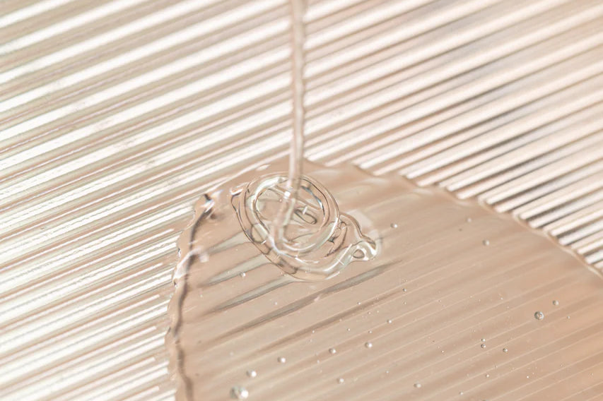 Oway Frequent Use Hair & Scalp Bath, a gentle daily shampoo for all hair types, is shown as a clear liquid poured onto a textured surface, with light reflecting off its sulfate-free formula and ridged grooves.