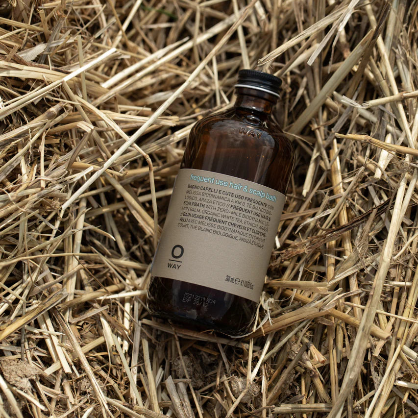 A brown glass bottle labeled “Oway Frequent Use Hair & Scalp Bath | Gentle Daily Shampoo for All Hair Types” by Oway rests on a bed of dry straw.