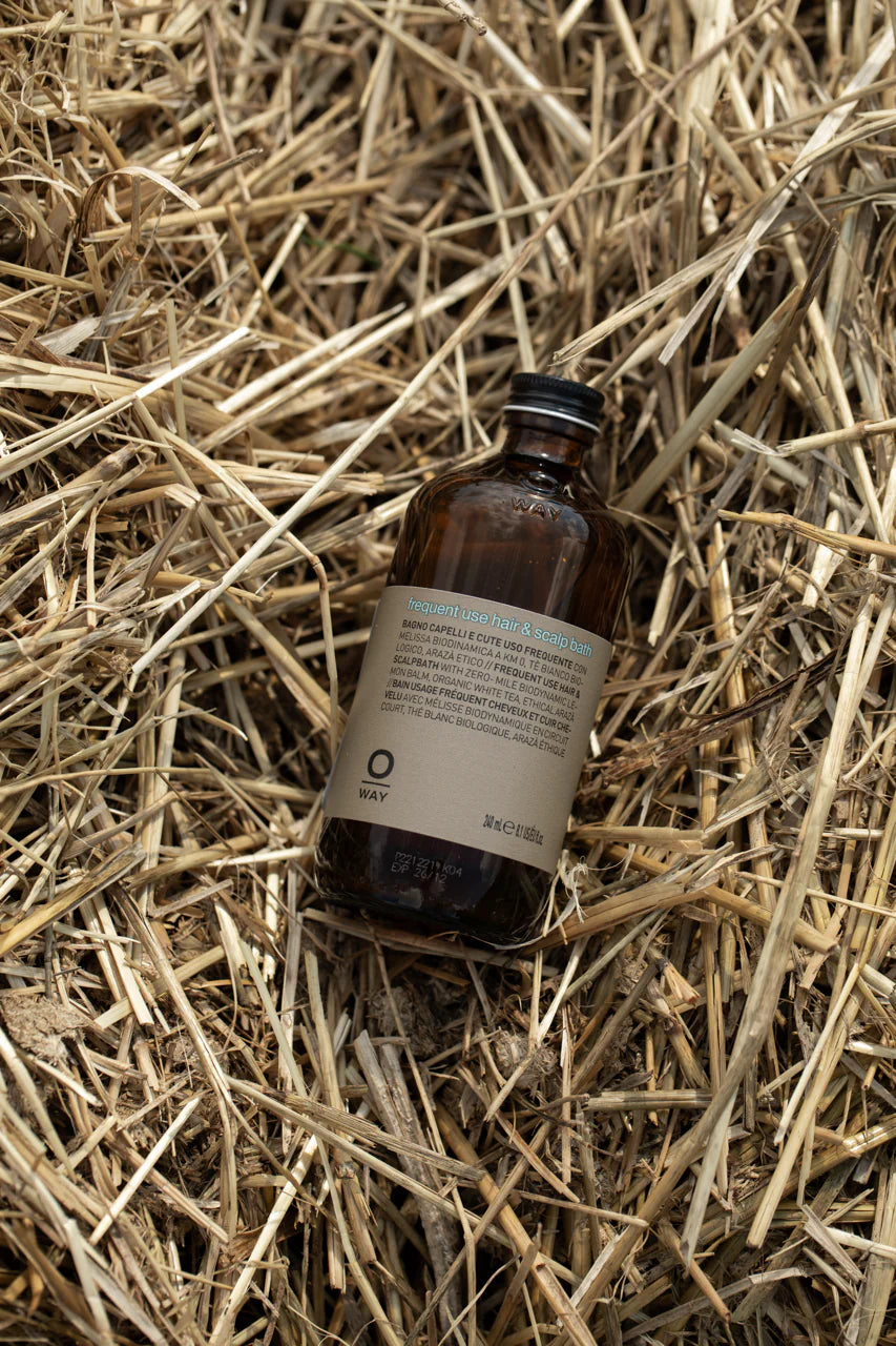 A brown glass bottle labeled “Oway Frequent Use Hair & Scalp Bath | Gentle Daily Shampoo for All Hair Types” by Oway rests on a bed of dry straw.