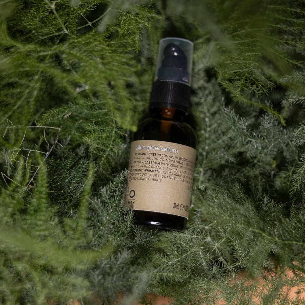 A brown glass bottle labeled "Oway Silk’N Glow Serum | Anti-Frizz Shine Serum for Dry, Frizzy Hair" with a black spray cap rests on lush green foliage.