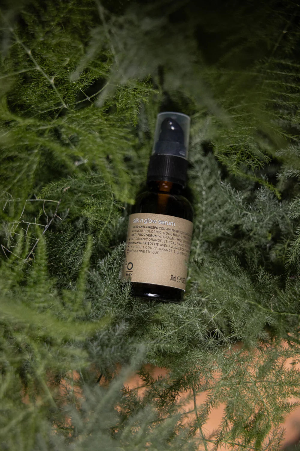 A brown glass bottle labeled "Oway Silk’N Glow Serum | Anti-Frizz Shine Serum for Dry, Frizzy Hair" with a black spray cap rests on lush green foliage.