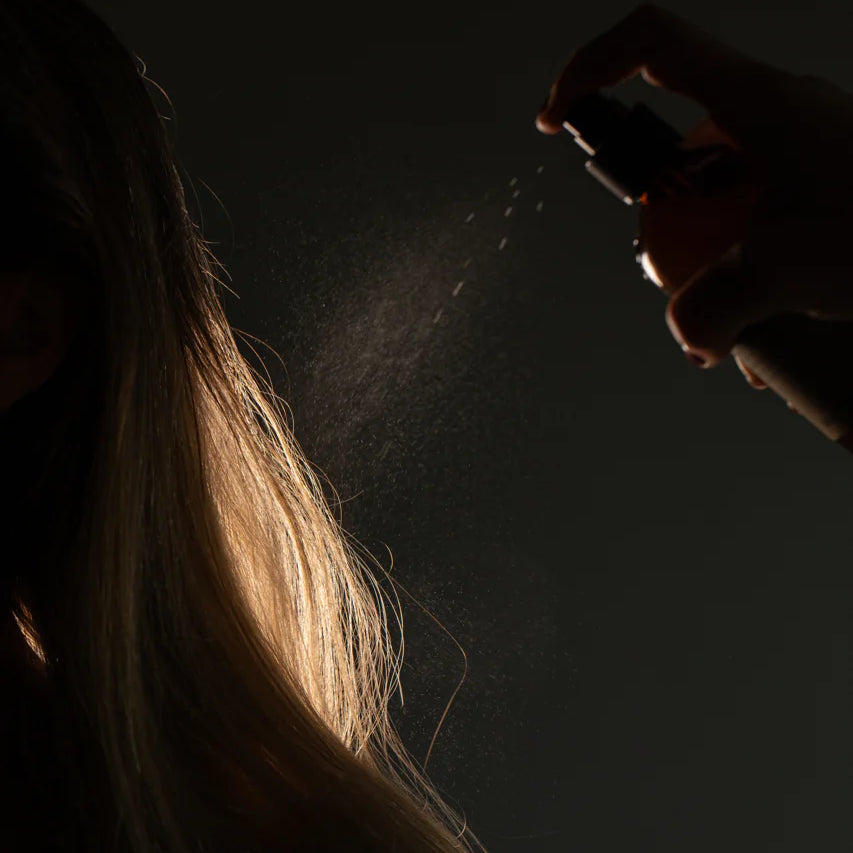 A hand sprays Oway Bodifying Hair Renewer onto long blonde hair, the mist infused with ethical marula oil illuminating fine strands against a dark background. This anti-hair fall densifying spray is ideal for fine hair.