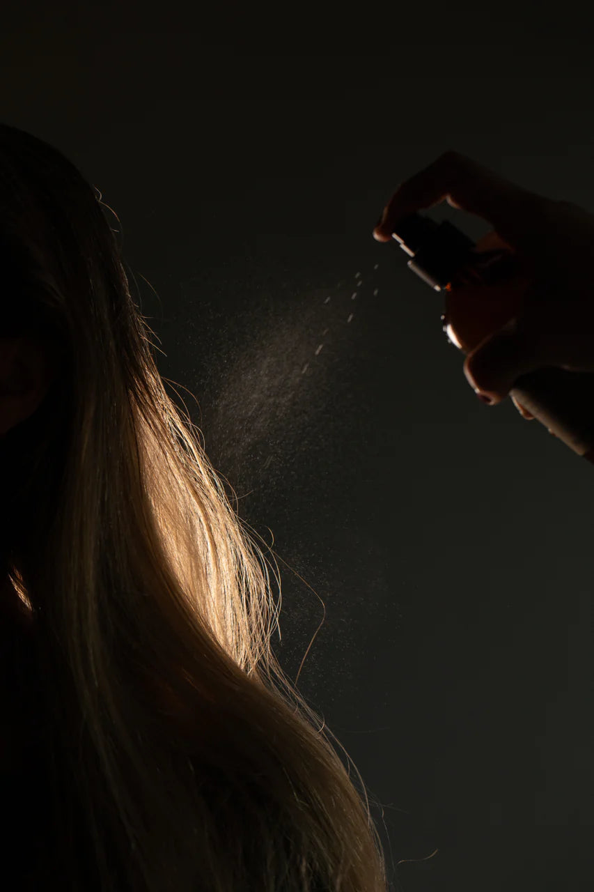 A hand sprays Oway Bodifying Hair Renewer onto long blonde hair, the mist infused with ethical marula oil illuminating fine strands against a dark background. This anti-hair fall densifying spray is ideal for fine hair.