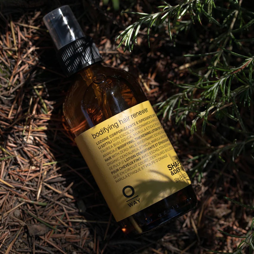 A brown Oway Bodifying Hair Renewer | Anti-Hair Fall Densifying Spray for Fine Hair bottle, enriched with ethical marula oil, sits among pine needles and green foliage.