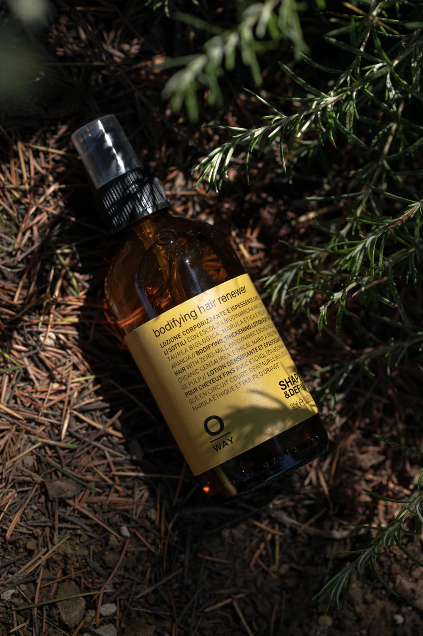 A brown Oway Bodifying Hair Renewer | Anti-Hair Fall Densifying Spray for Fine Hair bottle, enriched with ethical marula oil, sits among pine needles and green foliage.