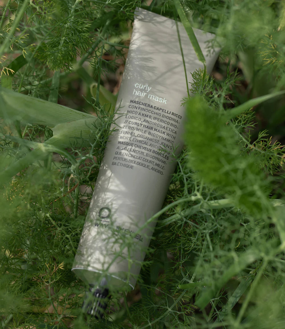 A tube of Oway Curly Hair Mask | Definition and Moisture Mask for Natural Curls rests among green leaves, its front label showing product details in multiple languages.