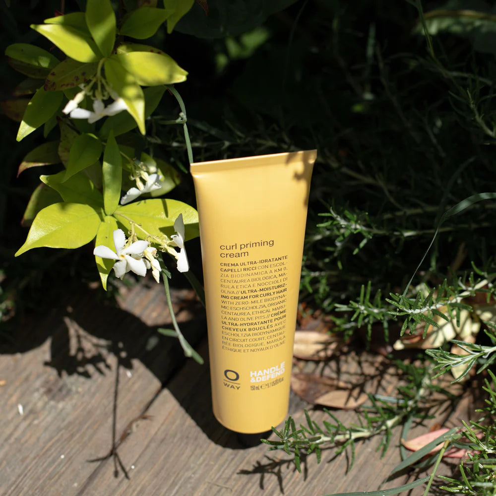 A tube of Oway Curl Priming Cream | Moisture-Rich Curl Primer for Frizz-Free Styling stands upright on wooden planks amid green foliage and white flowers, highlighting its promise of defined, nourished curls.