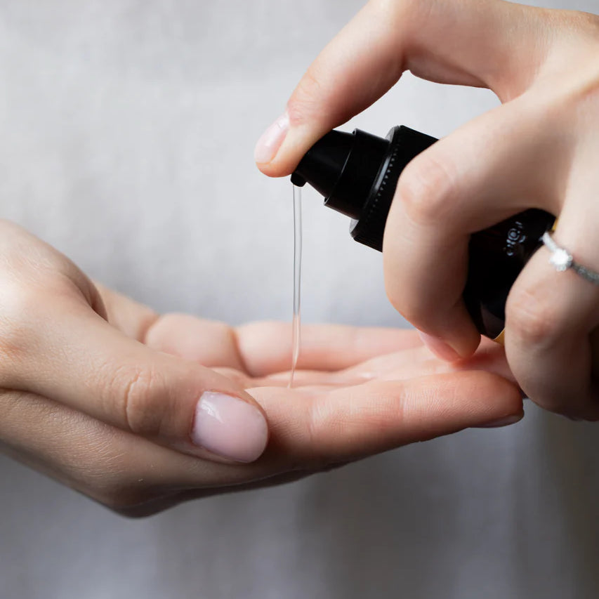 A person dispenses Oway Curly Potion | Curl-Defining Serum for Soft, Shiny Curls from a black bottle onto their palm, showing its frizz control and curl-enhancing benefits against a softly blurred background.