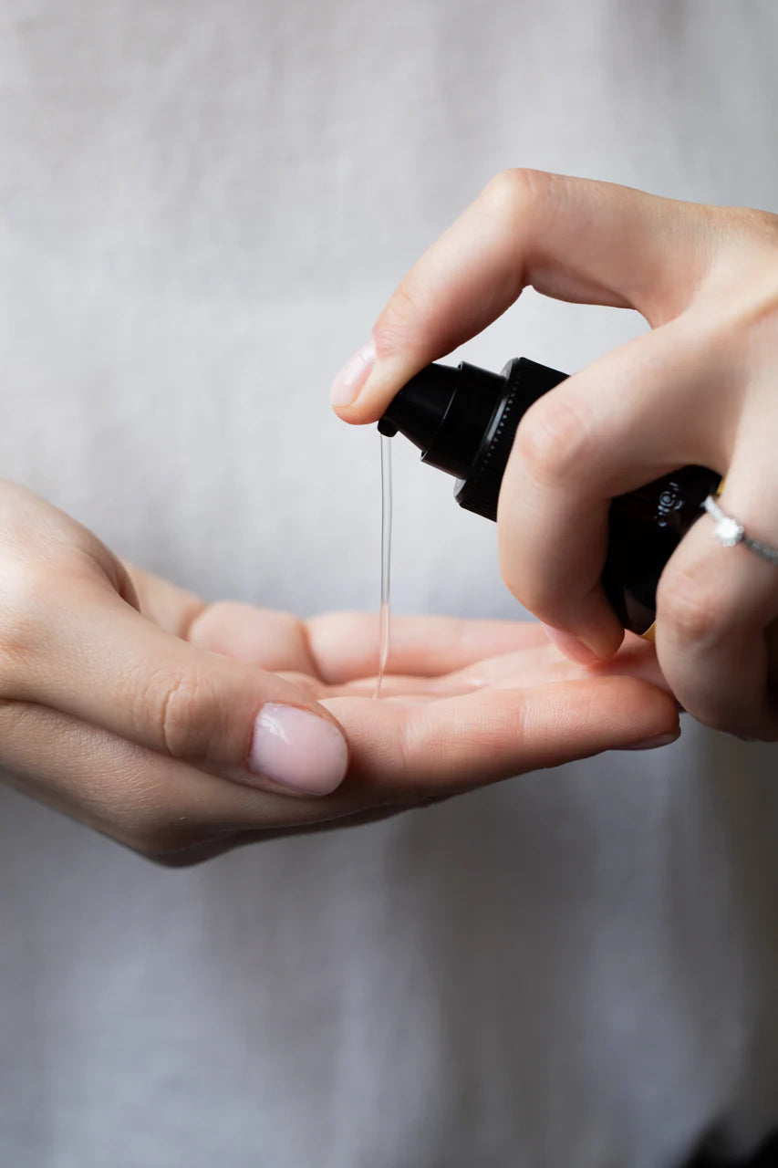 A person dispenses Oway Curly Potion | Curl-Defining Serum for Soft, Shiny Curls from a black bottle onto their palm, showing its frizz control and curl-enhancing benefits against a softly blurred background.