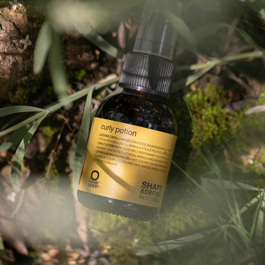 A bottle of Oway Curly Potion | Curl-Defining Serum for Soft, Shiny Curls rests on mossy ground among green grass and plants, highlighting the organic formula's natural connection and curl-enhancing benefits.