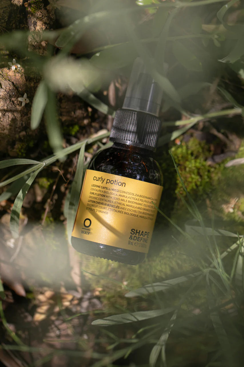 A bottle of Oway Curly Potion | Curl-Defining Serum for Soft, Shiny Curls rests on mossy ground among green grass and plants, highlighting the organic formula's natural connection and curl-enhancing benefits.