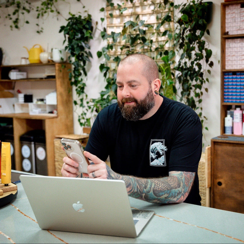A bearded man with tattoos sits at a desk, looking at his smartphone and smiling. An open laptop is in front of him. The background has shelves, plants, and various office supplies.