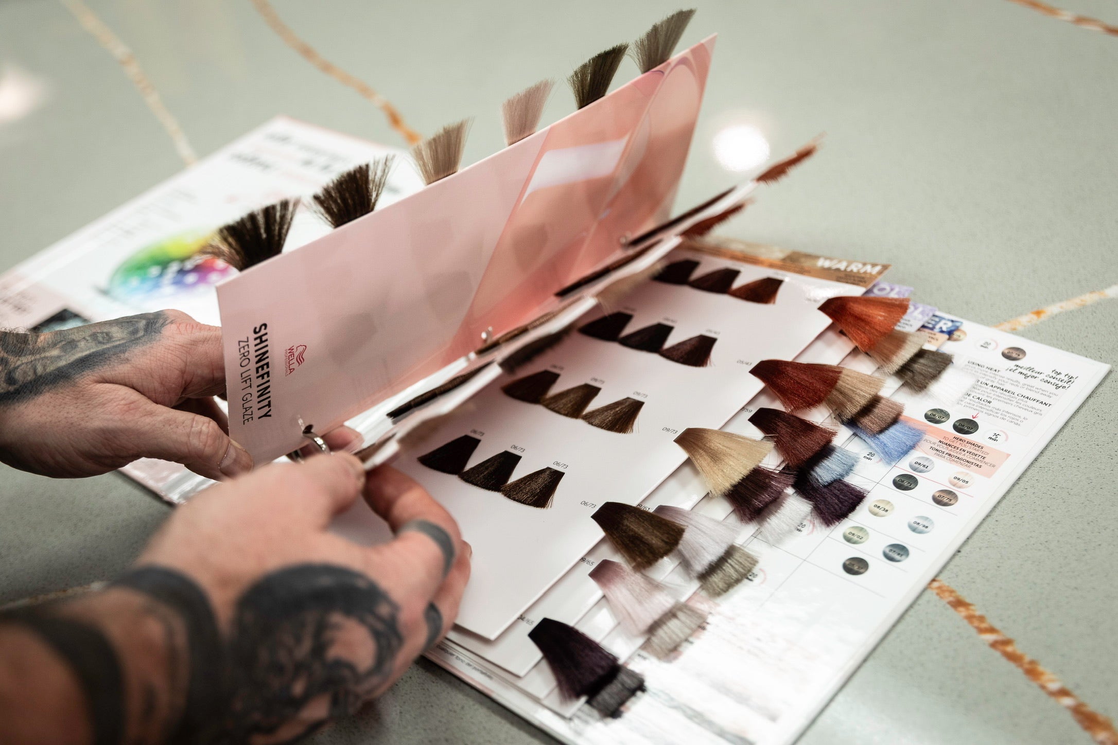 A person with tattooed hands flips through a hair color swatch book on a countertop, displaying a range of hair color samples from dark brown to blonde.