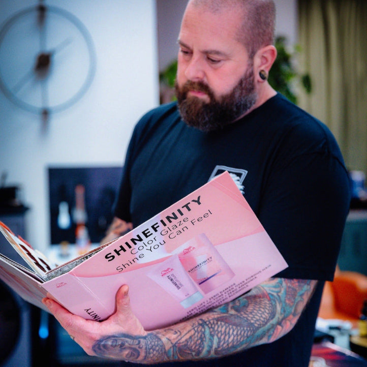 A bearded man with tattoos on his arms is standing indoors, wearing a black t-shirt, and reading a large booklet with the title "SHINEFINITY Color Glaze Shine You Can Feel" on the cover.