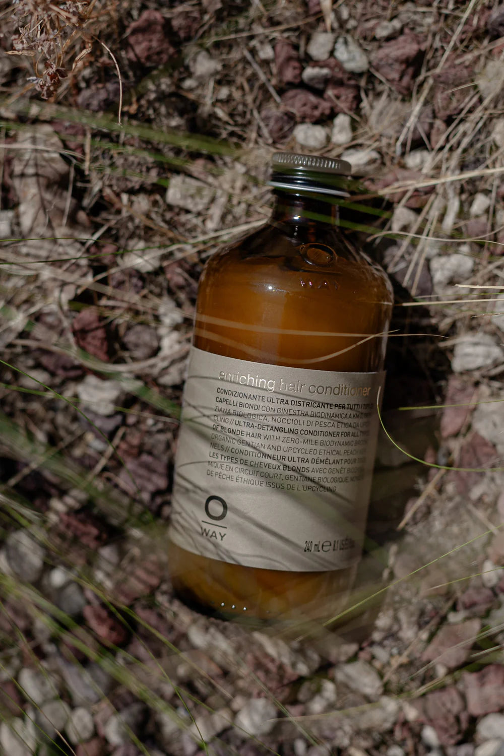 An Oway Enriching Hair Conditioner bottle for dry, brittle strands, featuring an ultra detangling plant-based formula, rests on rocky ground with dry grass.