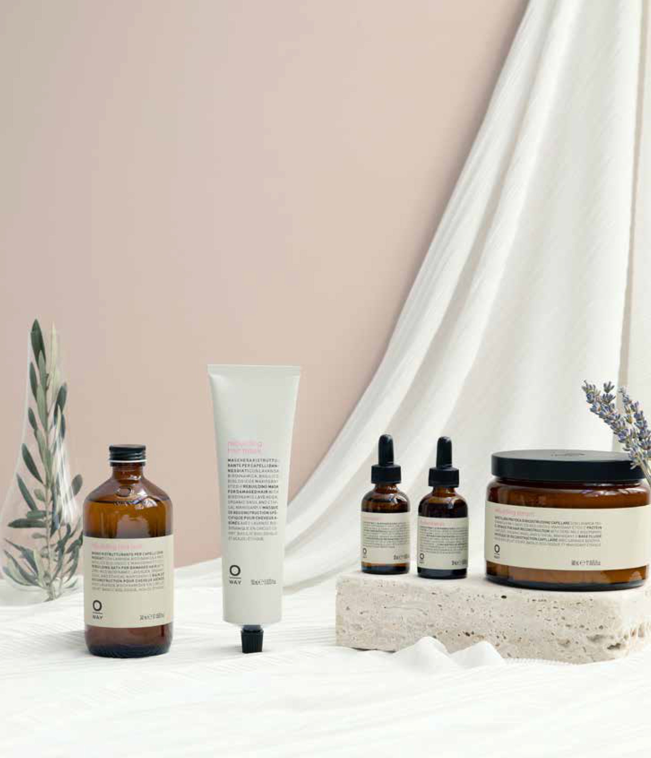 The Oway Rebuilding Hair Mask, a restorative mask for damage and instant repair, in amber packaging is displayed on light fabric with small plants and a textured stone.