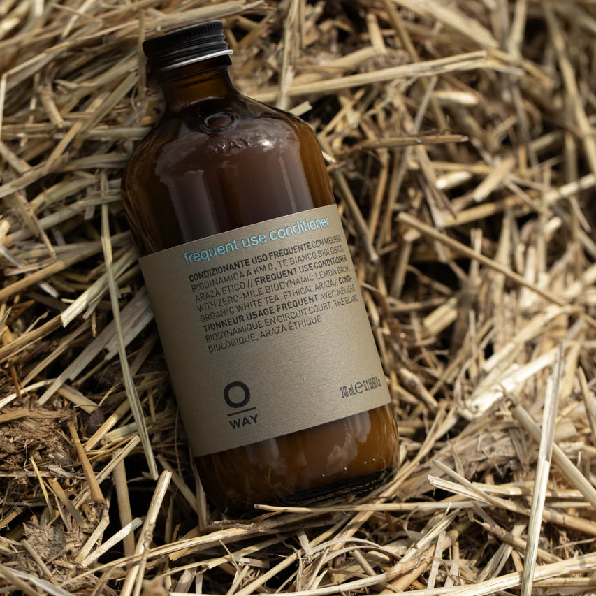 A brown glass bottle of Oway Frequent Use Conditioner | Lightweight Conditioner for Everyday Use rests on dry straw, displaying its label.