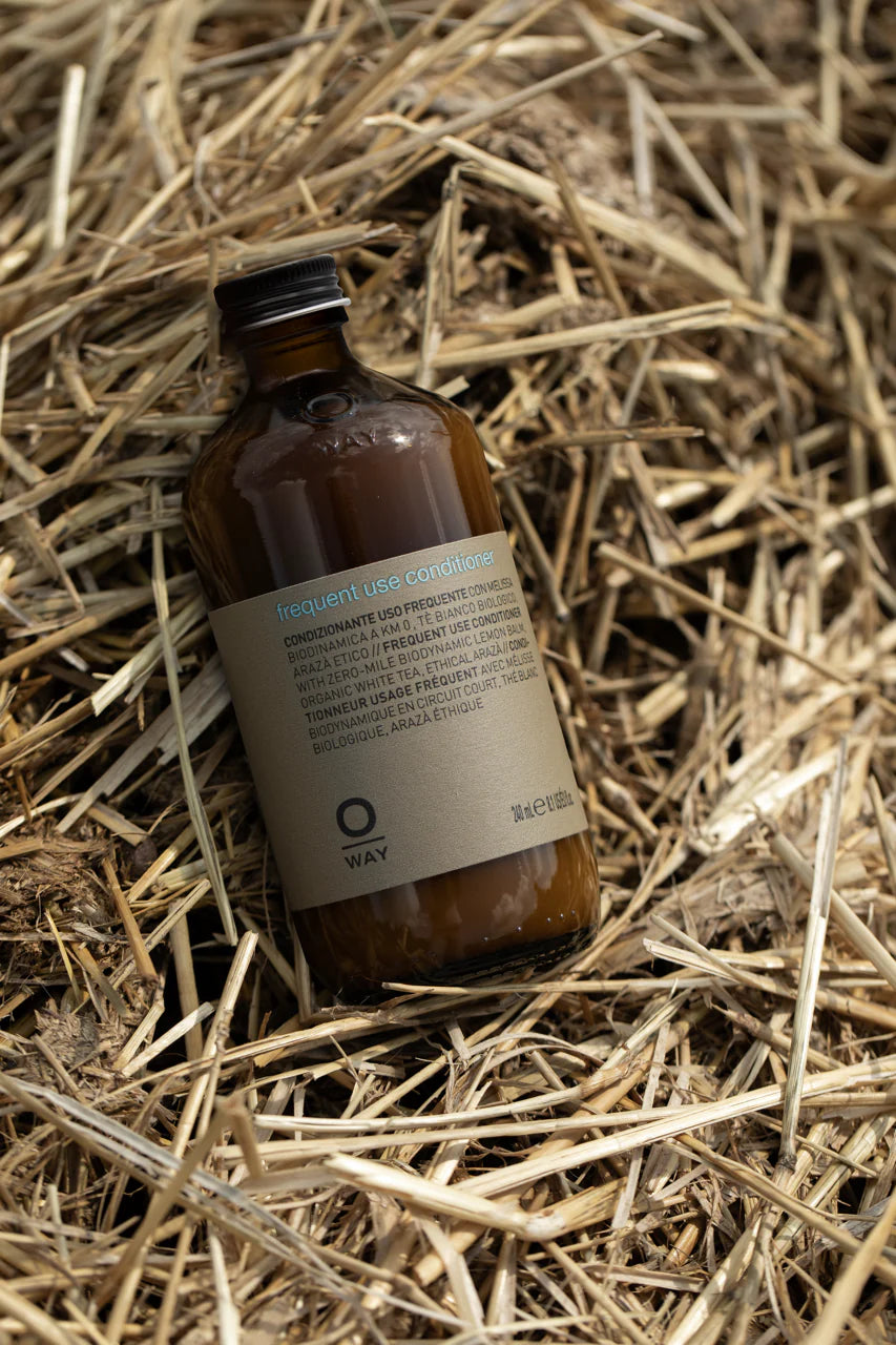 A brown glass bottle of Oway Frequent Use Conditioner | Lightweight Conditioner for Everyday Use rests on dry straw, displaying its label.