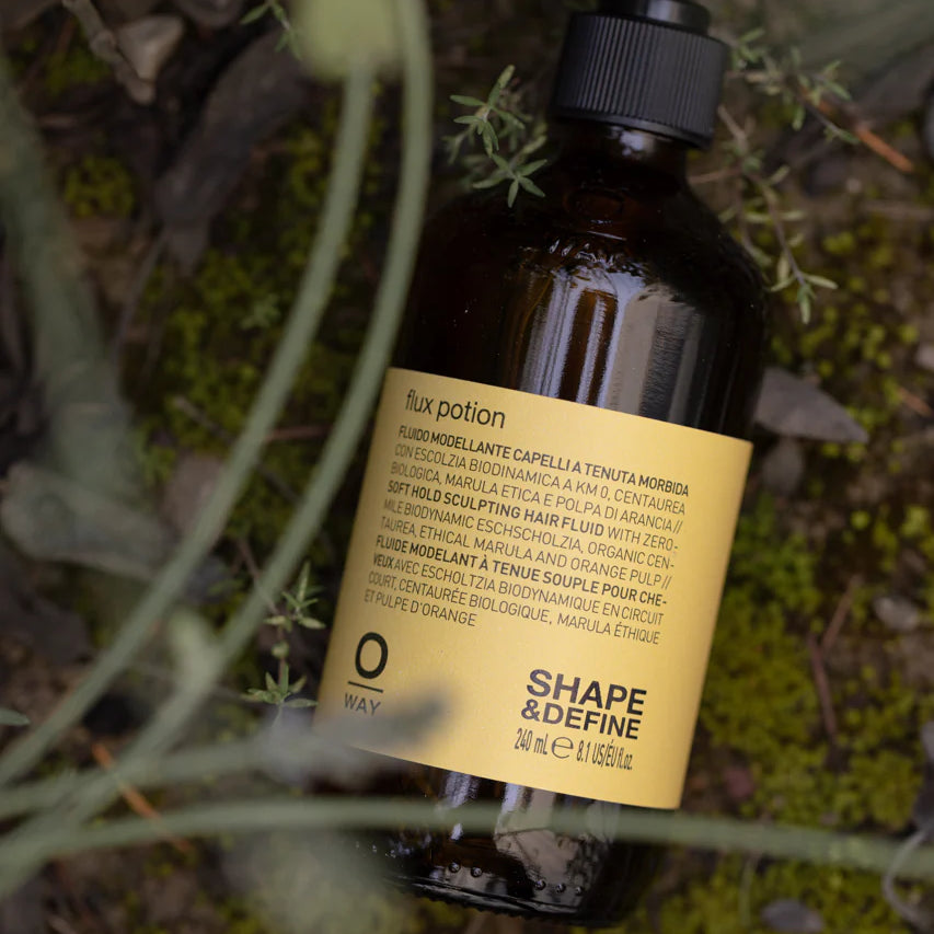 A brown bottle of Oway Flux Potion Lightweight Volumizing Serum for Soft Body and Hold, featuring a pump dispenser, sits among green plants outdoors.