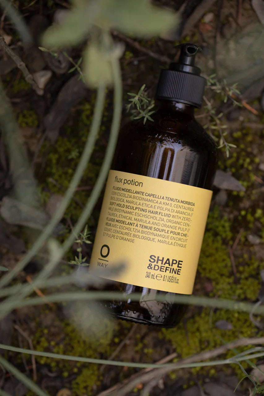 A brown bottle of Oway Flux Potion Lightweight Volumizing Serum for Soft Body and Hold, featuring a pump dispenser, sits among green plants outdoors.