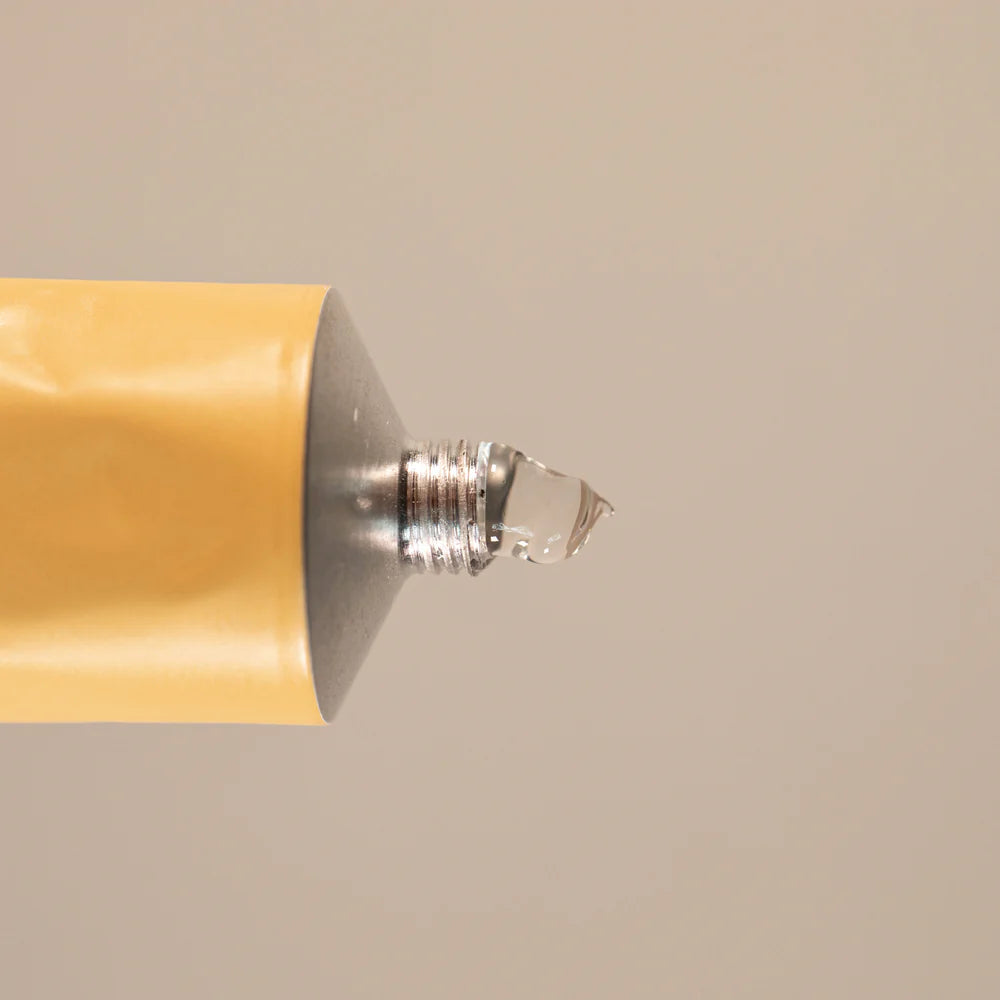 A close-up of Oway Glimmering Bond, a shine treatment for damaged, dull hair, shows clear gel enriched with ethical marula oil emerging from the open yellow tube against a plain beige background.