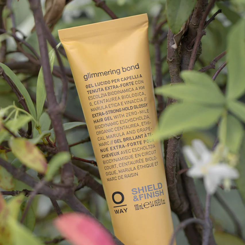 A yellow tube of Oway Glimmering Bond | Bonding Shine Treatment for Damaged, Dull Hair sits upright among green leaves, with a label spotlighting biodynamic botanicals and ethical marula oil for vibrant, lasting shine.
