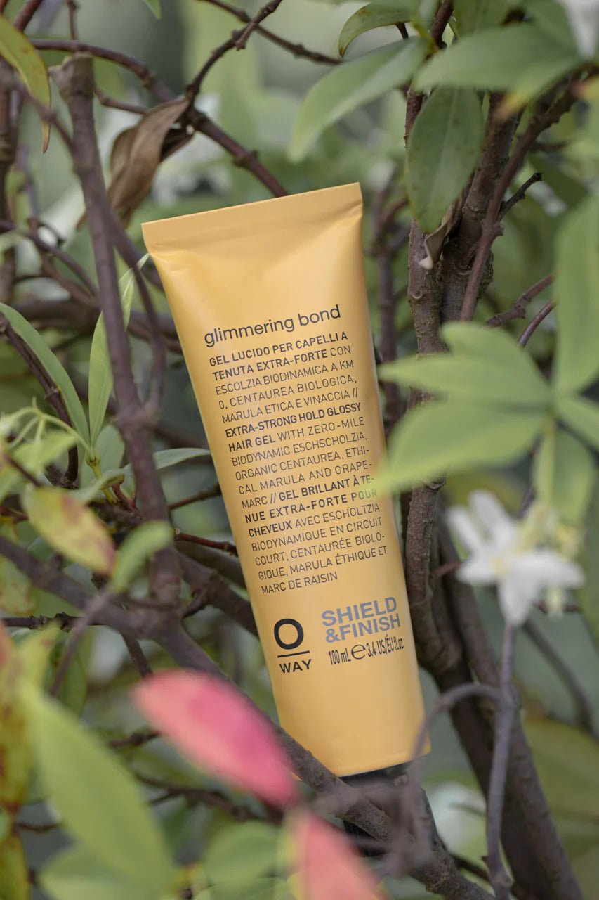 A yellow tube of Oway Glimmering Bond | Bonding Shine Treatment for Damaged, Dull Hair sits upright among green leaves, with a label spotlighting biodynamic botanicals and ethical marula oil for vibrant, lasting shine.