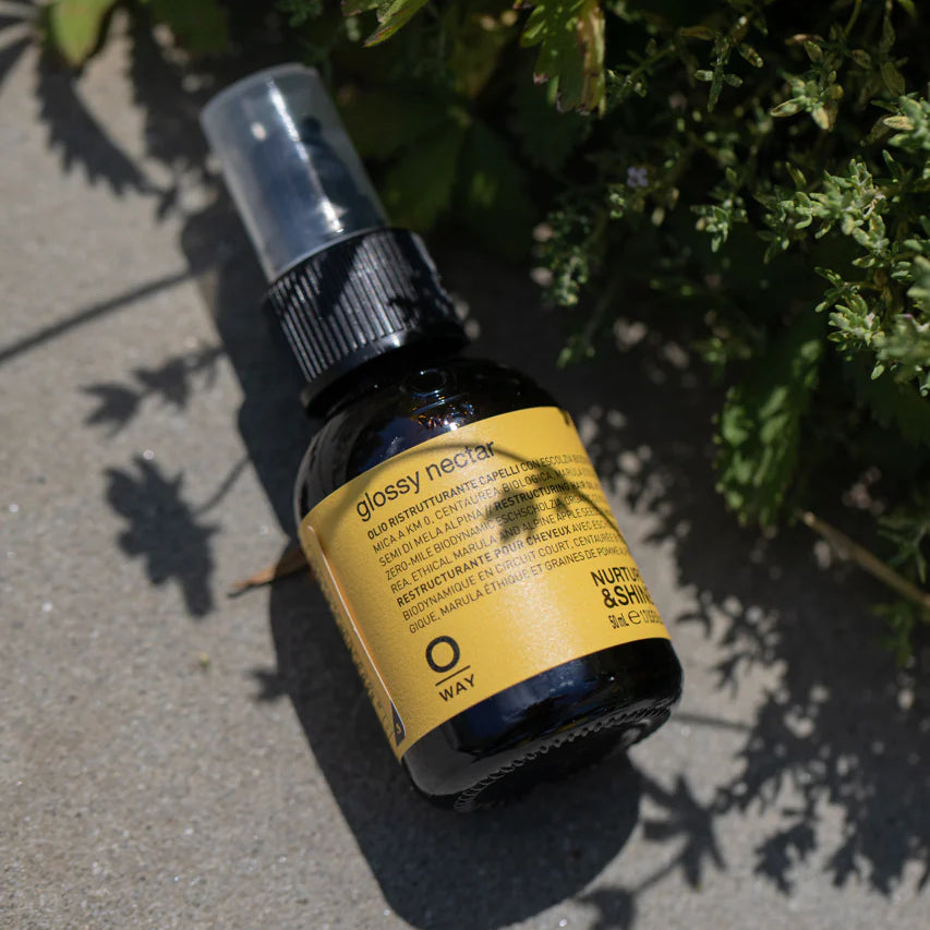 A small bottle of Oway Glossy Nectar | Rebuilding Oil Serum for Shine and Hair Strength rests on concrete in sunlight beside green leaves, offering frizz control, shine, and stronger hair.