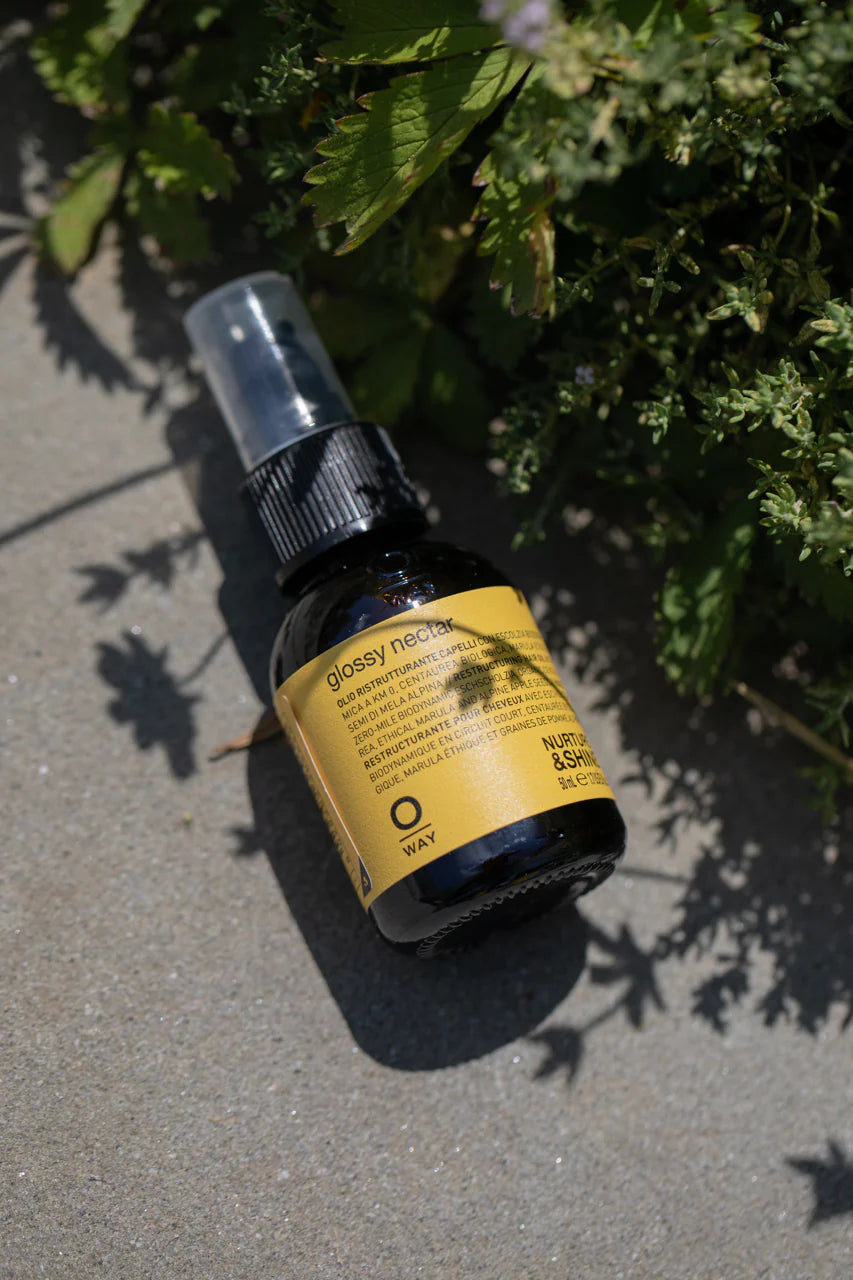 A small bottle of Oway Glossy Nectar | Rebuilding Oil Serum for Shine and Hair Strength rests on concrete in sunlight beside green leaves, offering frizz control, shine, and stronger hair.