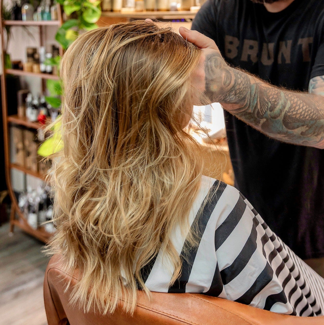 A person with long, wavy blonde hair sits in a salon chair wearing a striped cape while a tattooed hairstylist styles their hair. Shelves with hair products are visible in the background.