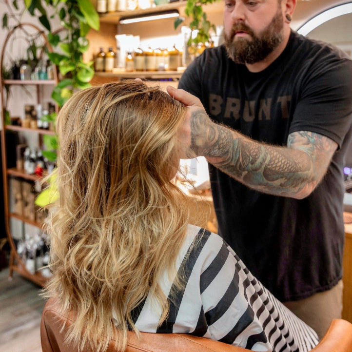 A person with long, wavy blonde hair sits in a salon chair wearing a striped cape, while a bearded hairstylist with tattoos styles their hair in a modern, well-lit salon with plants and shelves of products in the background.
