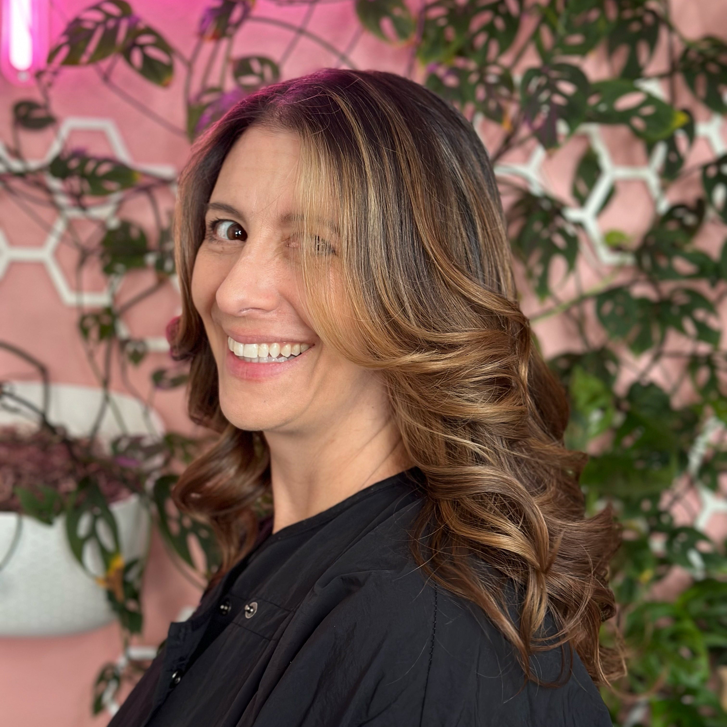 Woman with shoulder-length brown hair smiling in front of a pink wall with plants