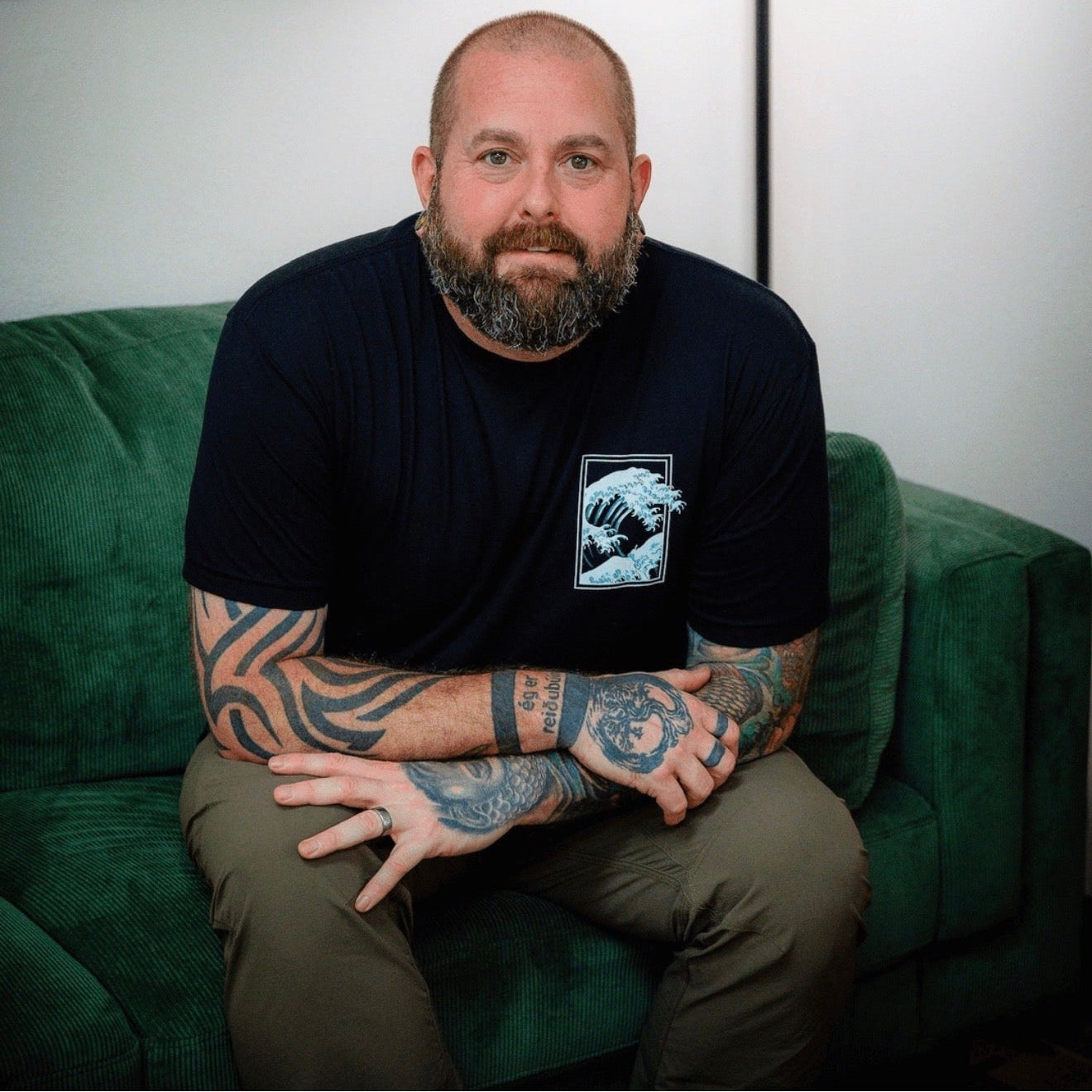 a man with tattoos, in a black shirt and green pants sitting on a green couch
