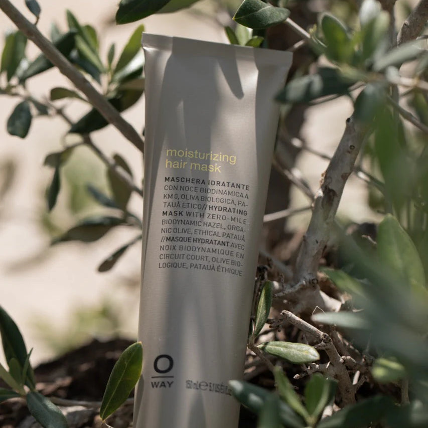 A tube of Oway Moisturizing Hair Mask rests among olive tree branches and leaves, its label visible—this deep hydration mask by Oway is ideal for dry or dehydrated hair.