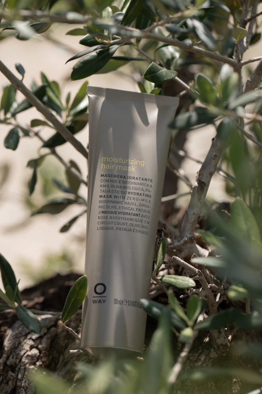 A tube of Oway Moisturizing Hair Mask rests among olive tree branches and leaves, its label visible—this deep hydration mask by Oway is ideal for dry or dehydrated hair.